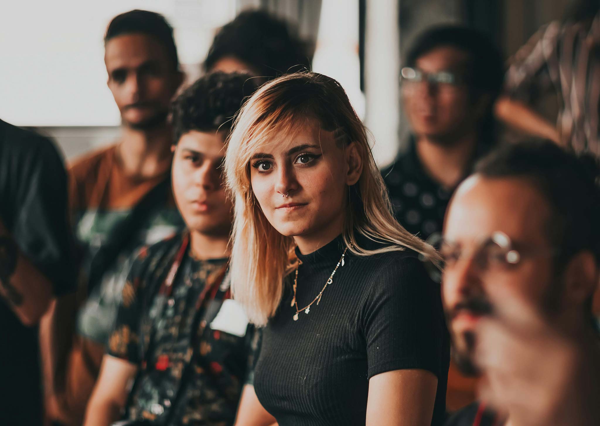 A photo of a crowded event, focussing on a single person stood centrally and listening to someone off camera